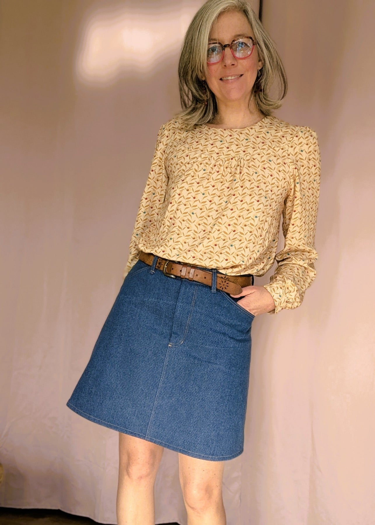 Jupe en jean bleu taille haute portée avec ceinture marron et blouse crème à motif, pose décontractée.
