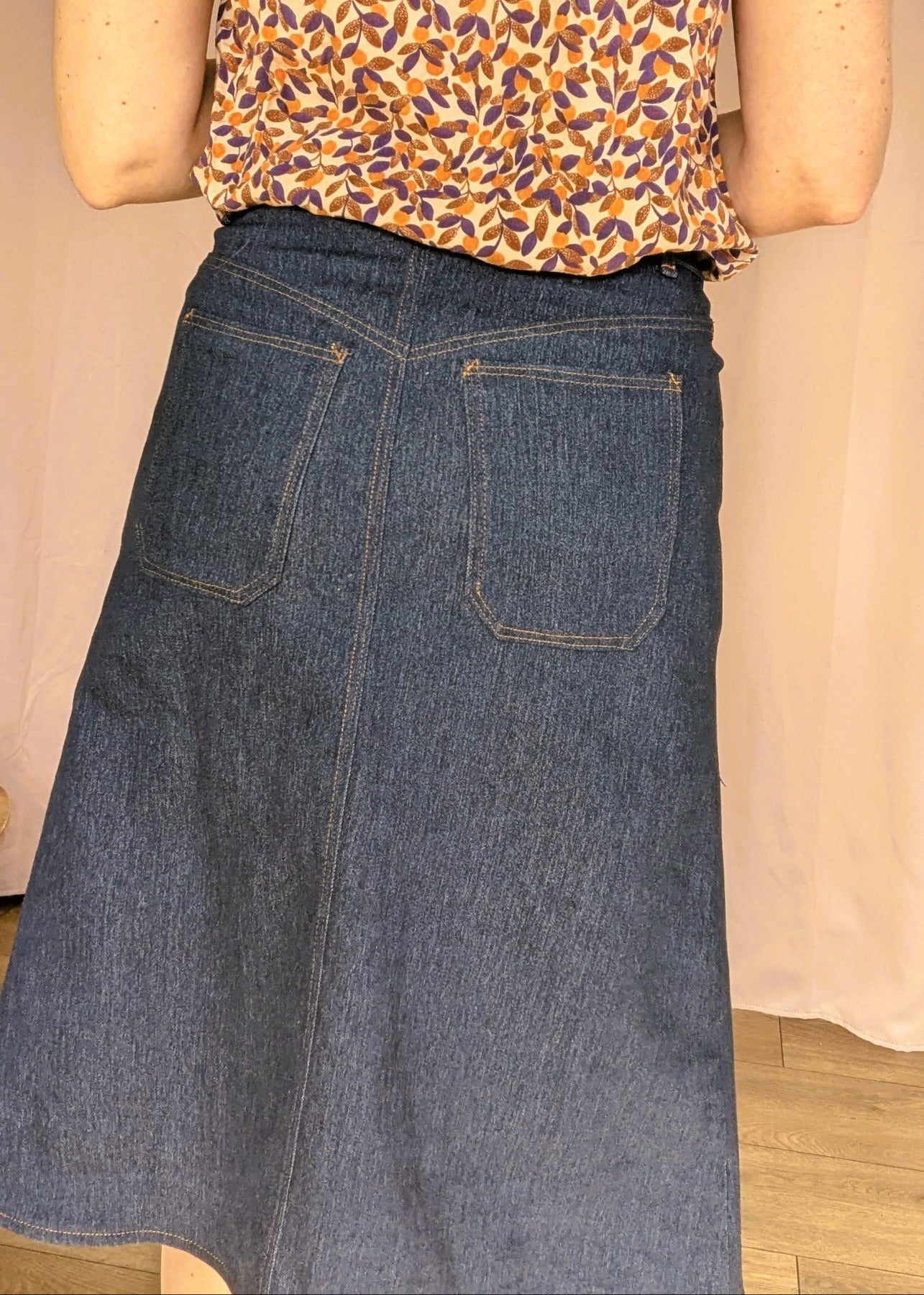 Jupe en jean taille moyenne vue de dos, avec poches arrière et couture apparente, associée à un top imprimé floral. La jupe Charmante est mise en valeur.