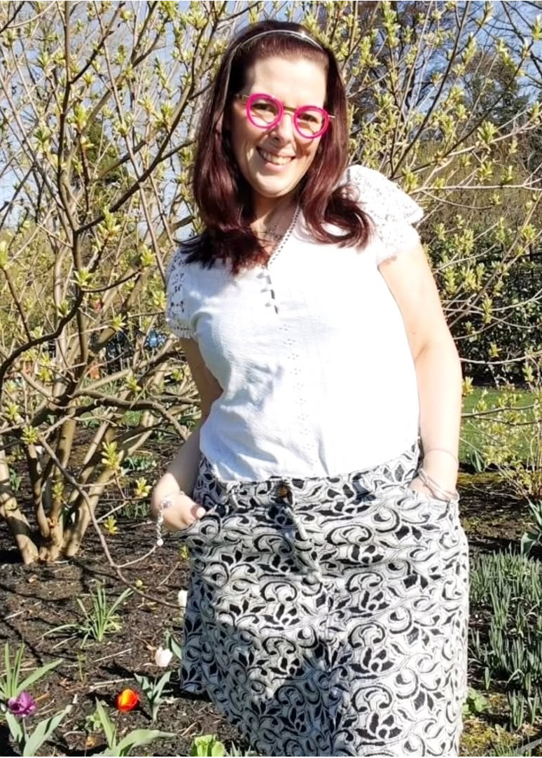Jupe Charmante en jean à motif écaille, portée par une femme souriante en chemise blanche, dans un jardin printanier. (La jupe est associée au produit)