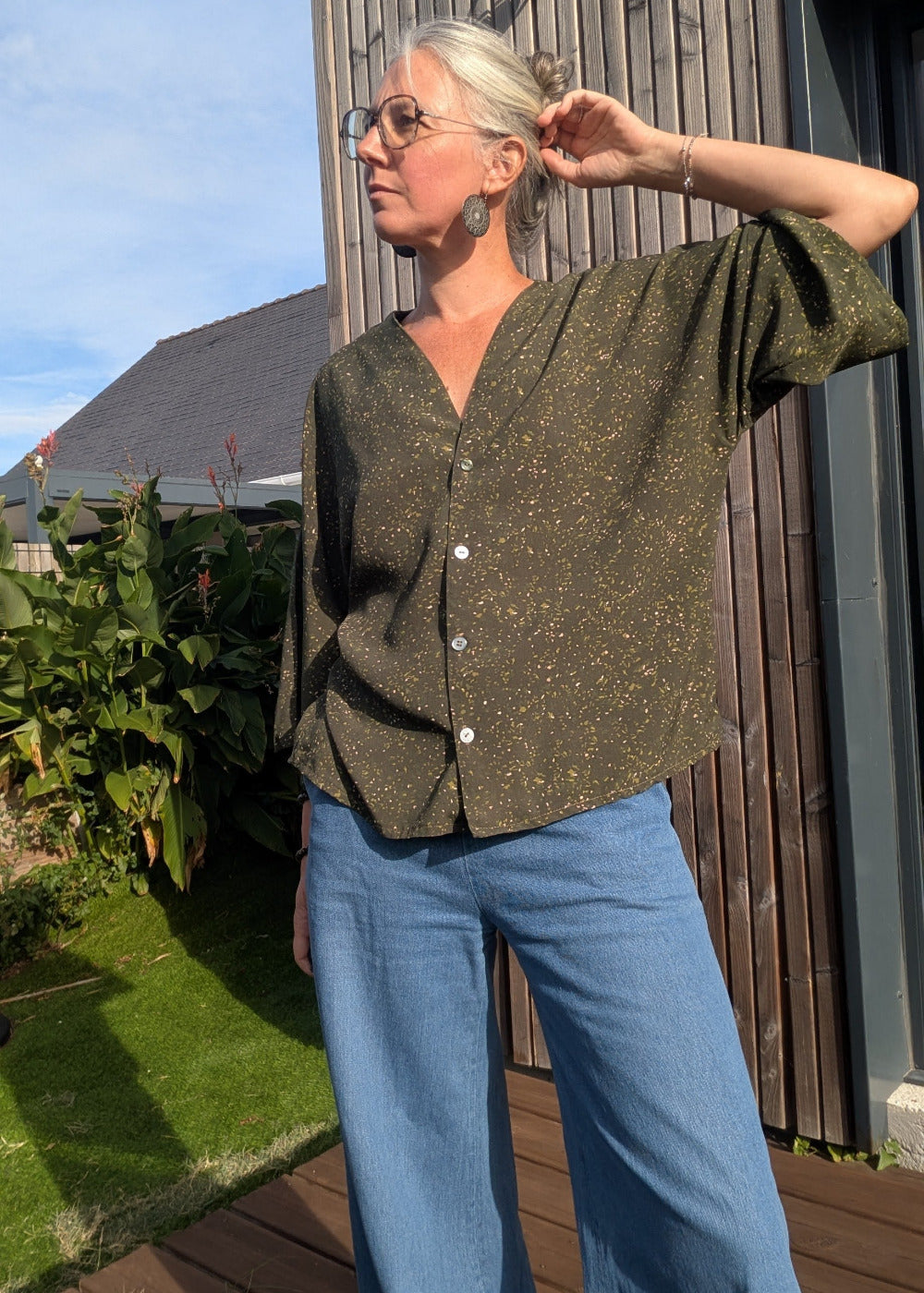 Femme âgée portant chemise olive à petites fleurs et jeans bleu, debout sur une terrasse en plein air, adjusting cheveux avec lunettes.
