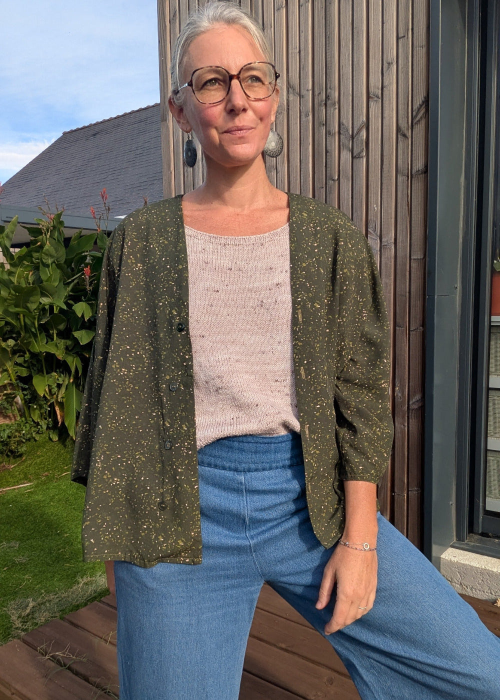 Homme/femme adulte portant veste kaki à petits pois dorés, pull beige, jeans bleus, lunettes rondes, bijoux pendants et posture dégingandée devant façade en bois. Keyword integration: si le mot-clé principal était « mode féminine éthique », on pourrait dire: tenue de mode féminine décontractée avec 