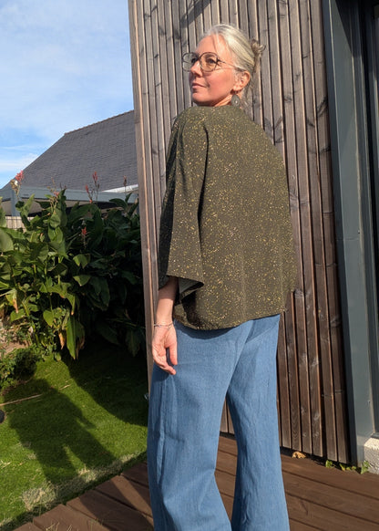 Blouse kaki à manches tres largas et finement à pois, jean bleu évasé, femme âgée aux cheveux gris et lunettes, debout sur terrasse en bois.