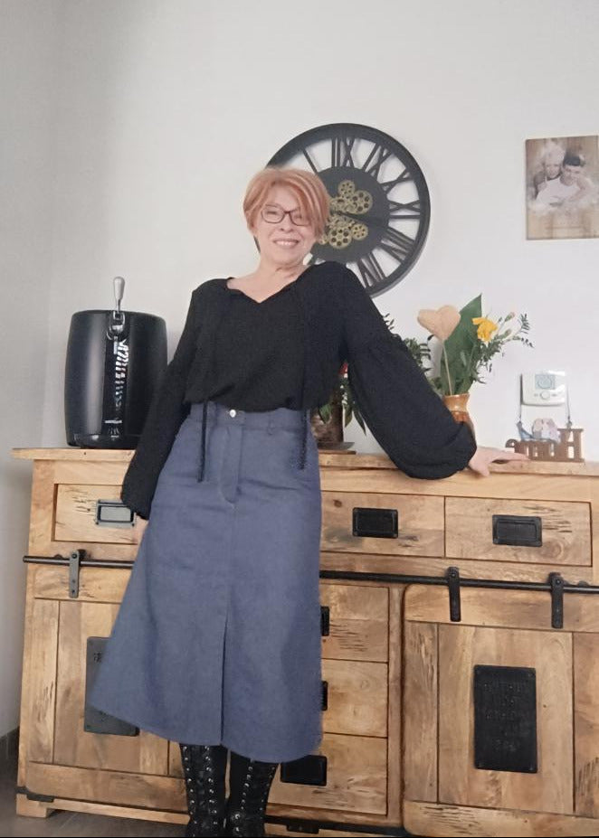 Jupe Charmante portée: une femme pose près d'une armoire en bois, portant une jupe bleu-gris et un haut noir à manches bouffantes.