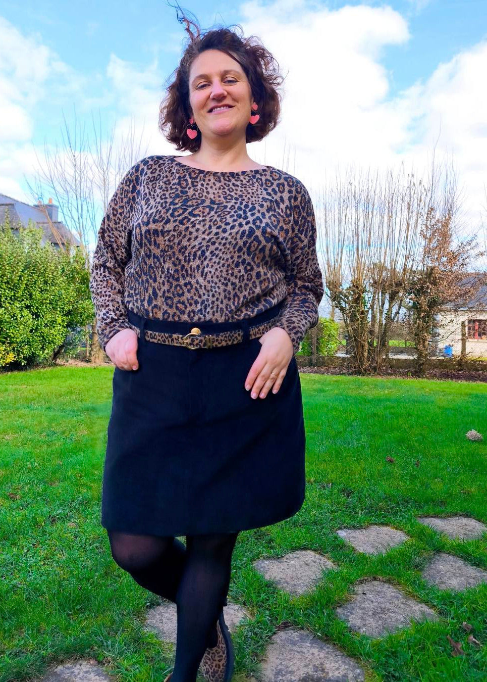 Jupe noire courte conifiée sur mannequin en plein air, blouse à imprimé léopard, ceinture hawk et collier rose.