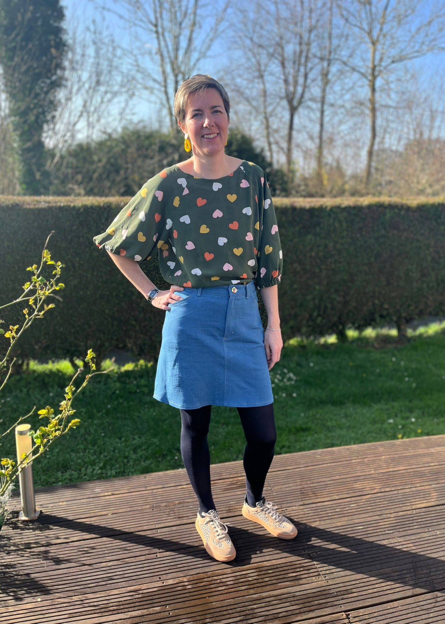 Jupe denim bleue A-line avec motif de cœurs, mancherons ondulés et look printanier. Contenu: modèle posant dehors, t-shirt à cœurs, collants noirs et sneakers. La jupe Charmante présentée avec tuto patron.