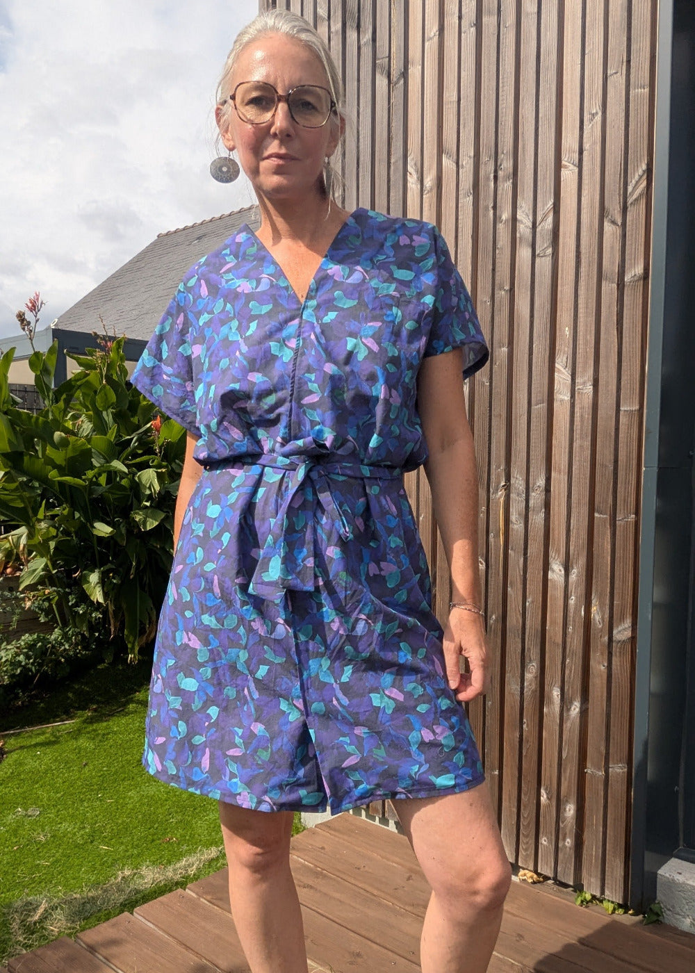 Personne portant une robe-short bleu à motifs verts, ceinturée à la taille avec nœud, lunettes rondes et grand bouclé d’oreille, debout sur une terrasse en bois près d’un mur en bois.