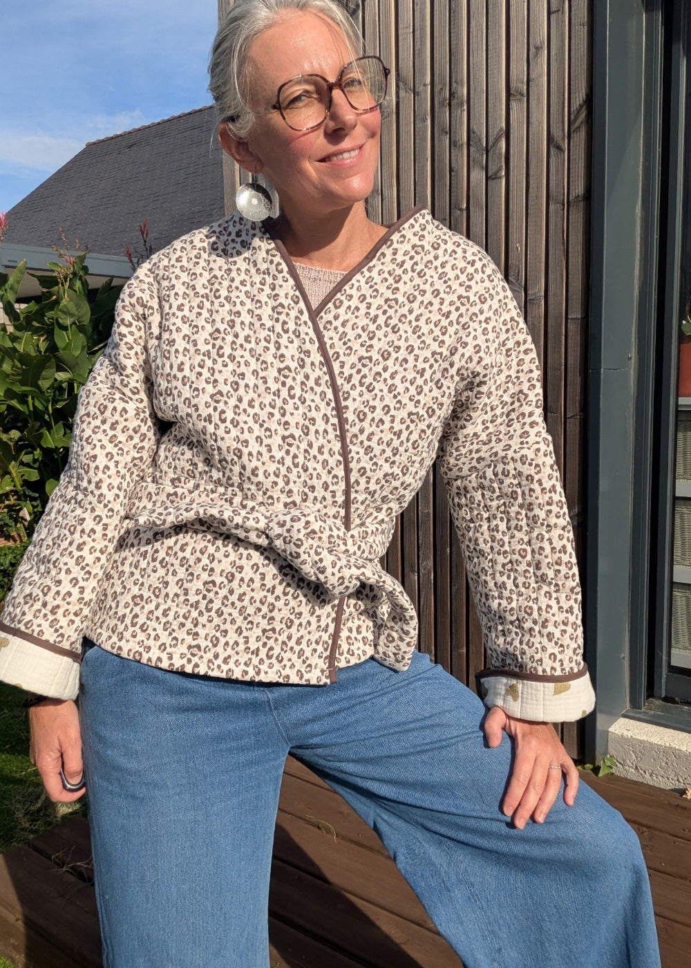 Mujer portant veste imprimé léopard, nœud à la taille, jean bleu, lunettes rondes, pose décontractée sur terrasse.
