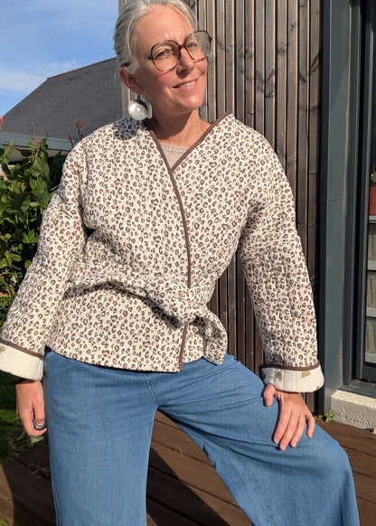 Mujer portant veste imprimé léopard, nœud à la taille, jean bleu, lunettes rondes, pose décontractée sur terrasse.
