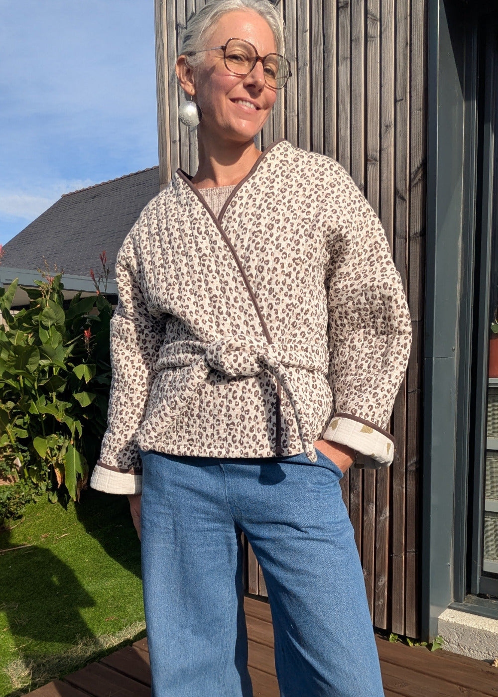 Vêtement décontracté: femme debout en plein soleil, veste Kimono à motif léopard beige, ceinture à la taille, manches retroussées, jean bleu clair et grandes lunettes.