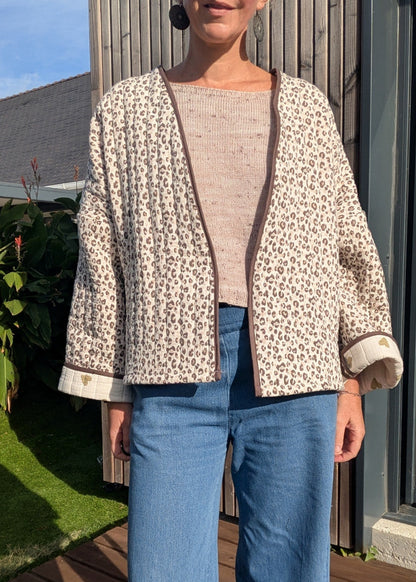 Veste kimono beige à imprimé léopard, revers et ourlets brun clair; pull beige en maille, jeans bleu clair, et boucles d’oreilles noires visibles.