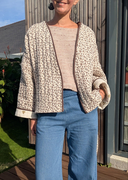 Personne debout dehors porte une veste beige à motif léopard, manches retroussées, sur un pull rosé clair, jeans bleus.