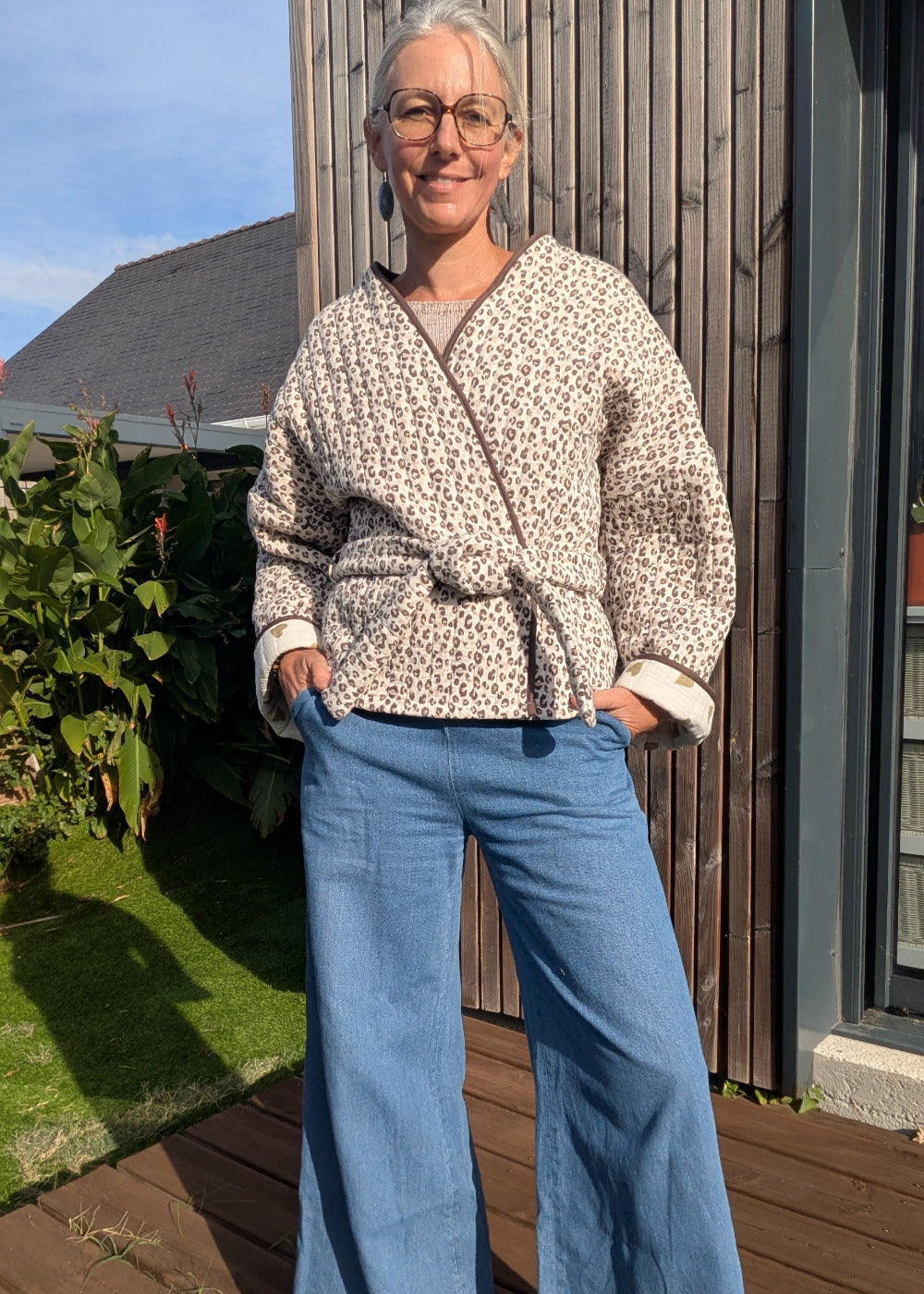 Homme ou femme debout en extérieur, veste kimono à imprimé léopard, ceinture à la taille, jeans bleu clair, lunettes, sourire, fond bois et jardin.