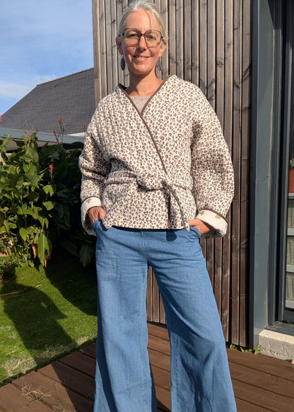 Homme ou femme debout en extérieur, veste kimono à imprimé léopard, ceinture à la taille, jeans bleu clair, lunettes, sourire, fond bois et jardin.