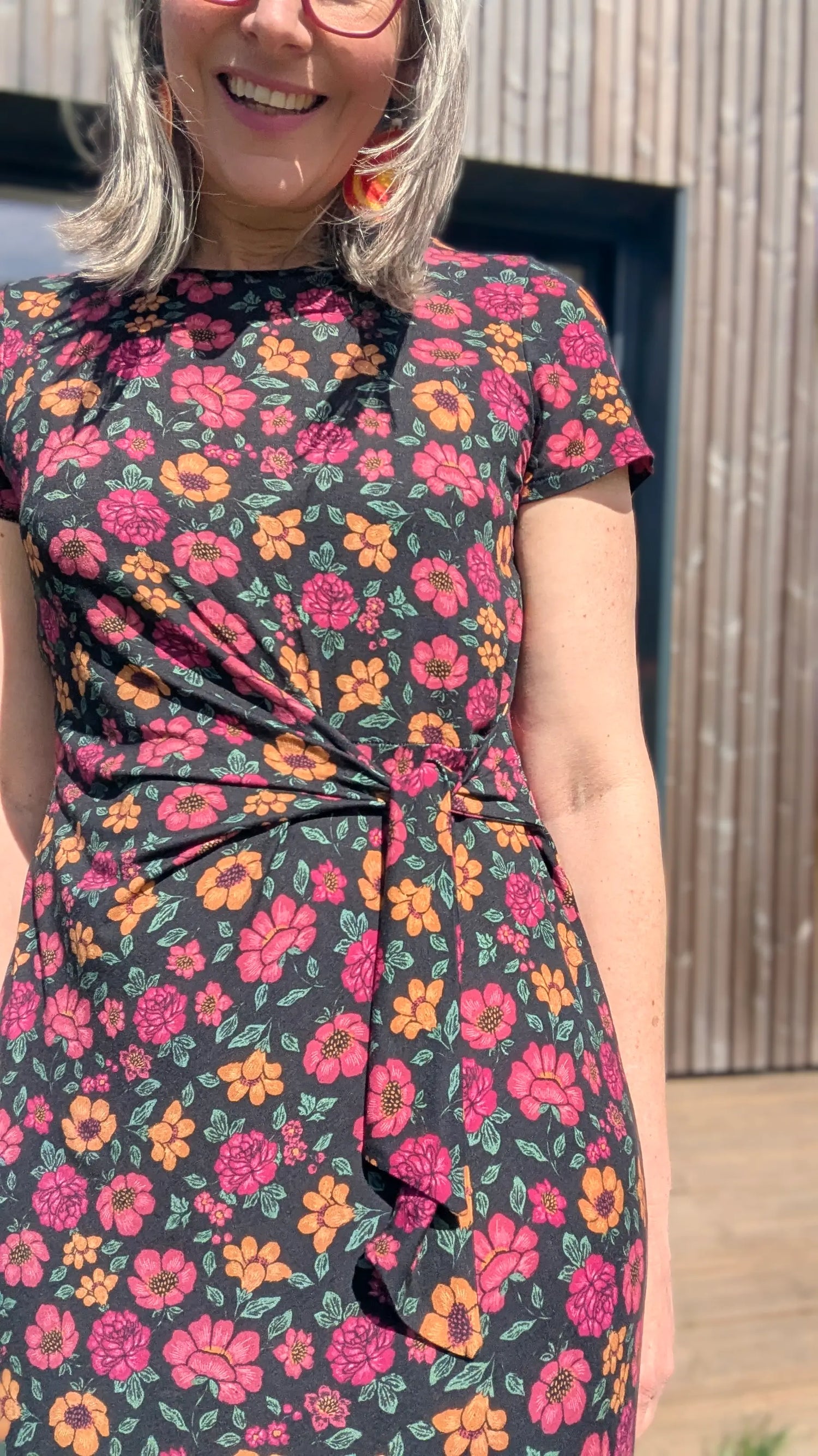 Je femme souriante porte une robe à motifs floraux roses et orange sur fond noir, cintrée à la taille avec nouage sur le devant.