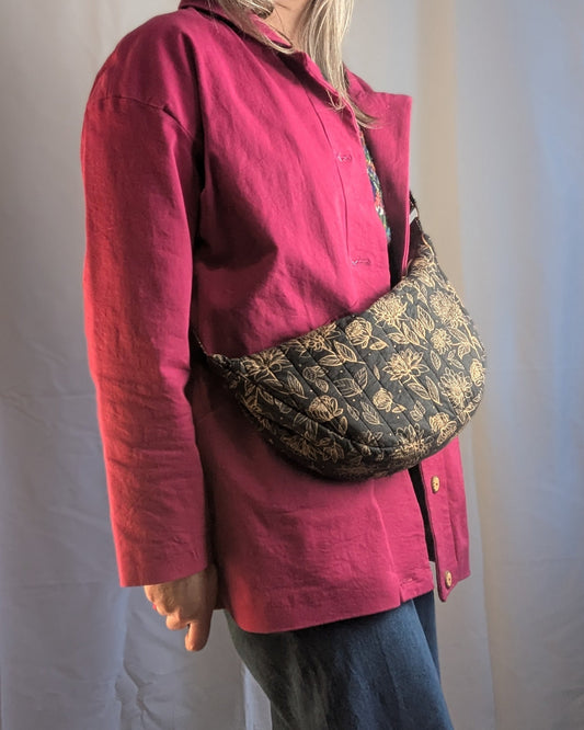 Sacoche en bandoulière noire à motif floral doré portée sur l’épaule d’une personne portant une chemise magenta et jean. Signature de l’ensemble de patrons couture - Chac Demi-lune et Chac Cabas.